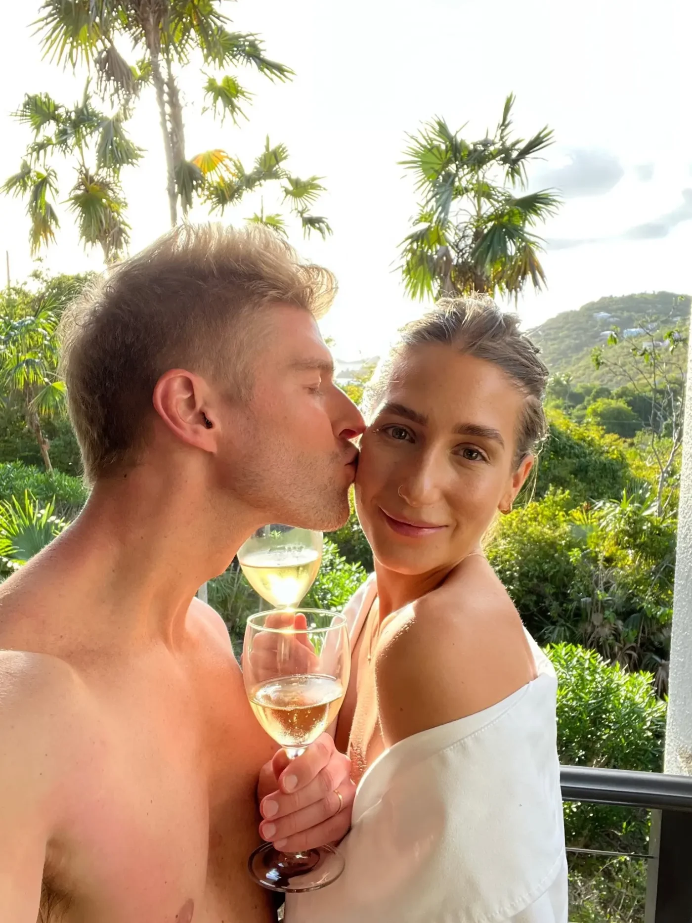 Kyle Cooke kissing Amanda Batula on the cheek while holding wine glasses on their honeymoon.