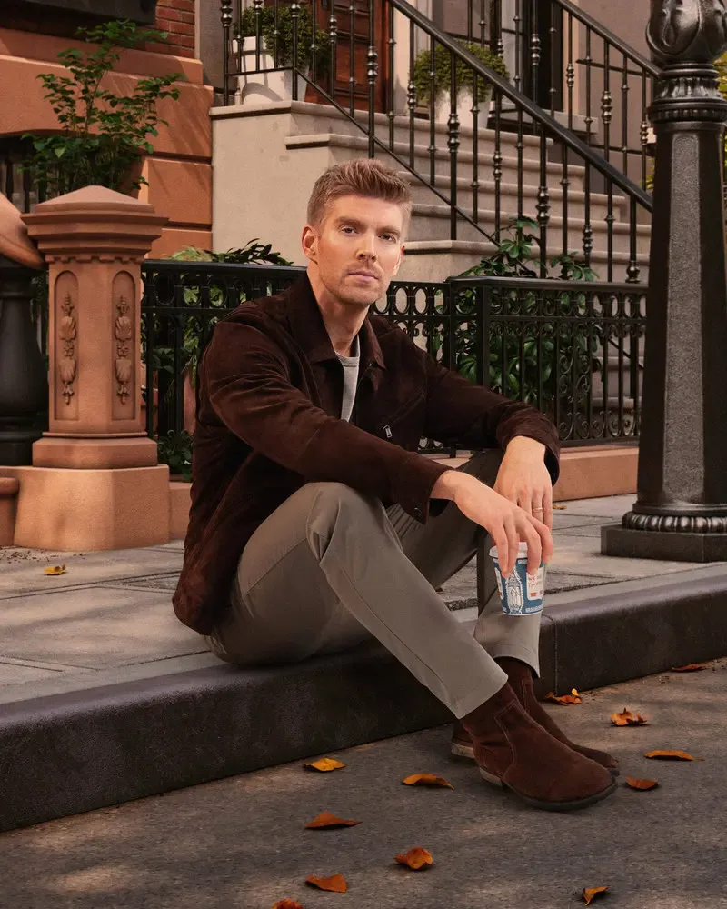Kyle Cooke sitting on a city sidewalk, wearing a brown jacket, holding a to-go coffee cup.