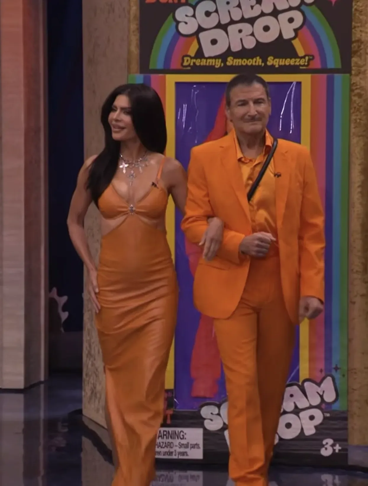 Lisa Rinna and Michael Gelman walk arm-in-arm across a stage with a multi-colored background. They are both dressed in head-to-toe orange outfits, and Rinna is wearing a long brunette wig to resemble Kylie Jenner.