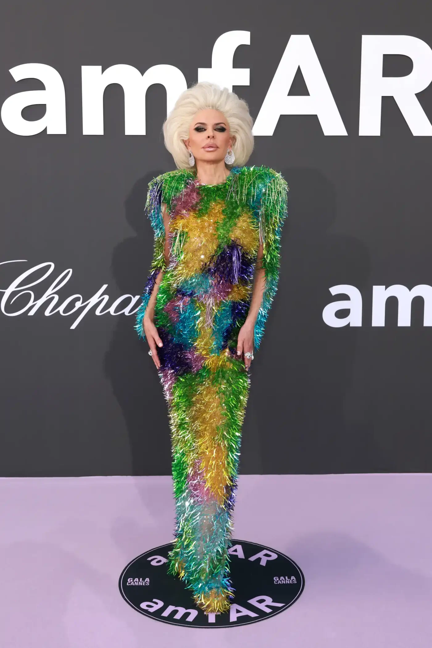 Lisa rinna posses on the red carpet in a platinum-blonde wig styled as a shoulder-grazing blowout. She is wearing a dramatic, colorful evening gown covered in glittering green, purple, yellow, blue, and pink fringe.