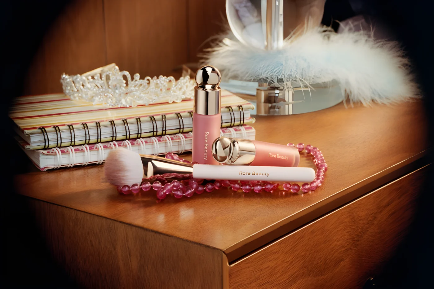 Makeup products by Rare Beauty on a wooden surface surrounded by notebooks, a tiara, and decorative beads.