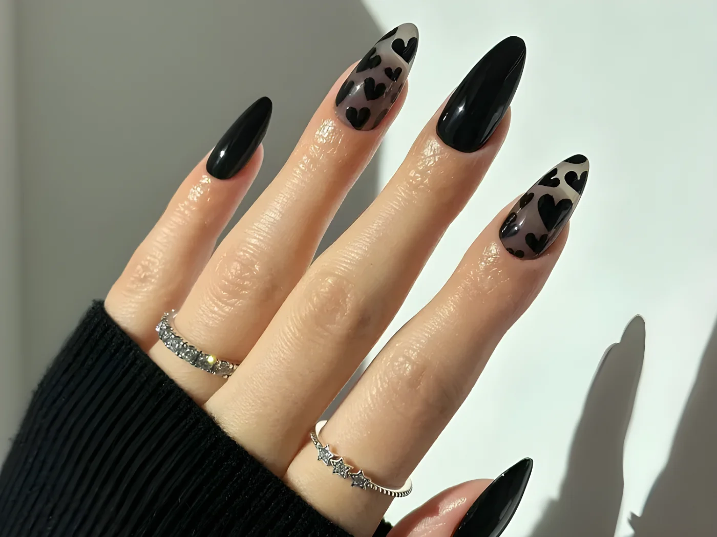 Manicured hand with black nails and heart designs on two fingers wearing silver rings and dark clothing