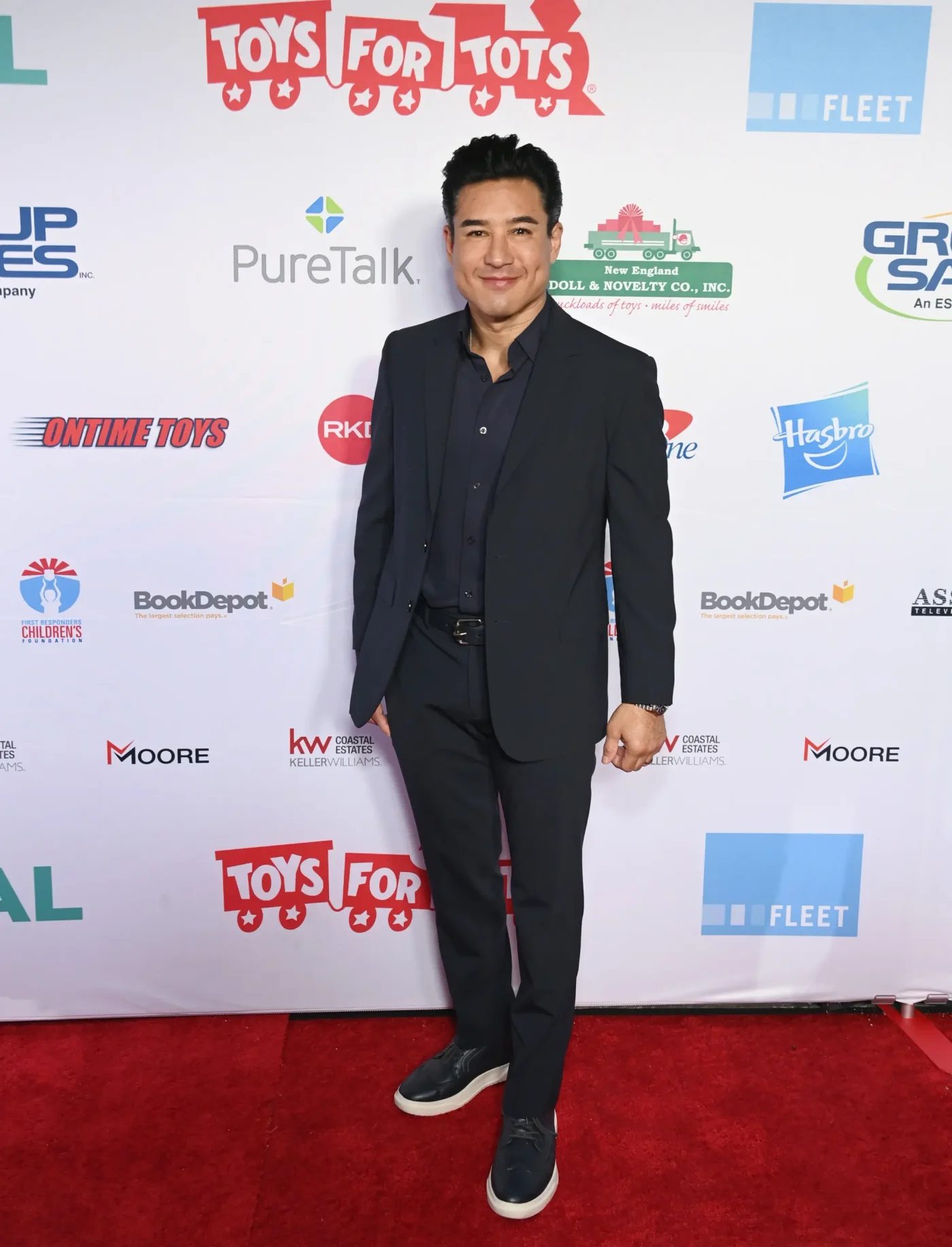 Mario Lopez poses in a dark suit on a red carpet in front of a sponsor backdrop.