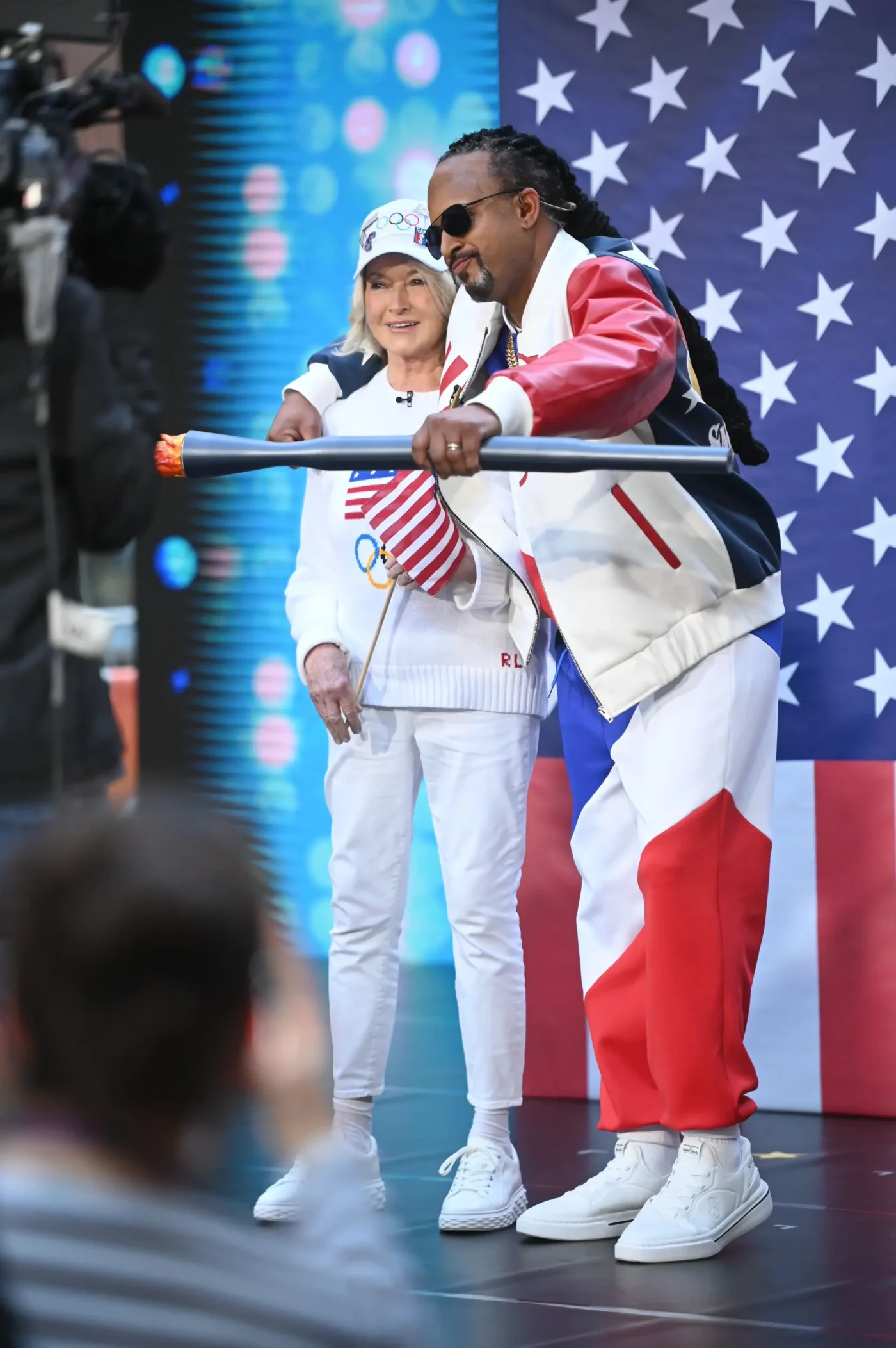 Martha Stewart and Craig Melvin dressed as Snoop Dogg and Martha Stewart holding an Olympic torch in front of an American flag backdrop.