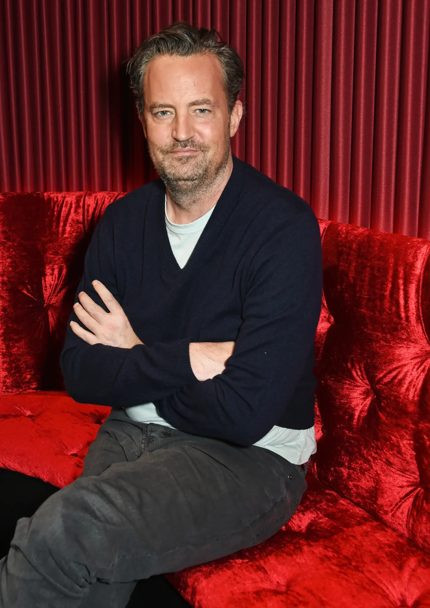 Matthew Perry sitting on a red velvet couch with his arms crossed during a photocall for his play