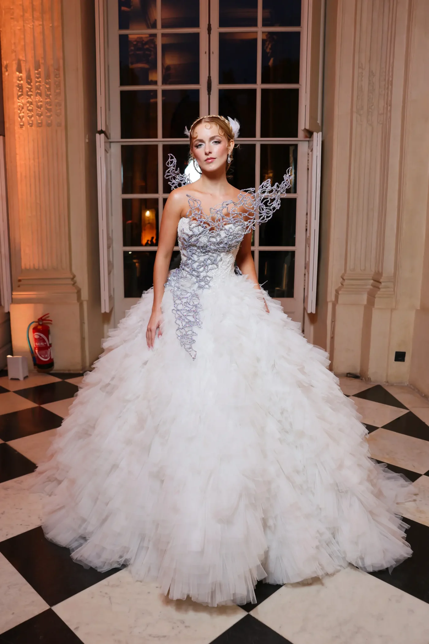 Mckenna Grace stands in a grand room, wearing a voluminous white ballgown adorned with intricate silver filigree.