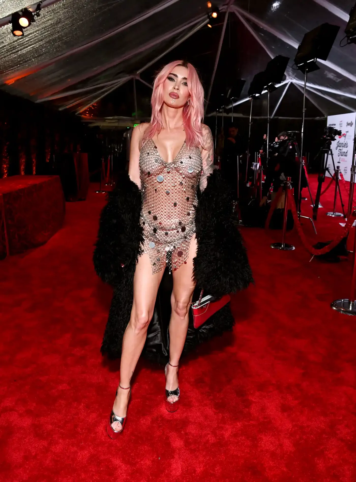 Megan Fox on a red carpet while wearing a short, see-through chain dress and black fur coat