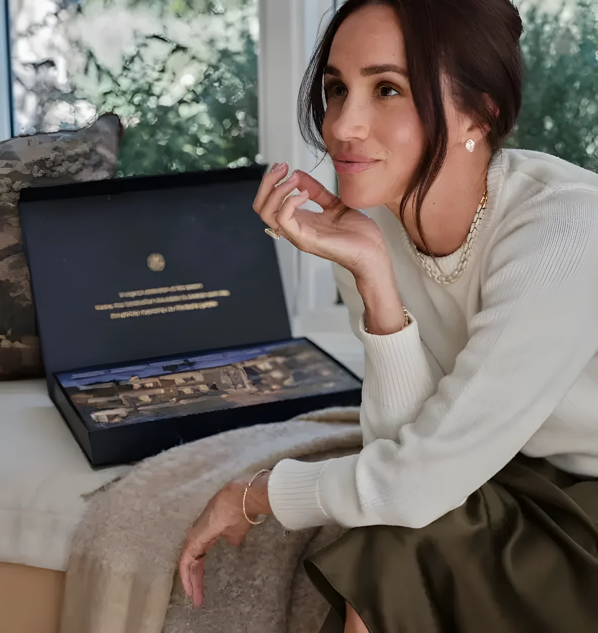 Meghan, Duchess of Sussex, wearing a white sweater and green skirt, sitting on a couch with a black box beside her.