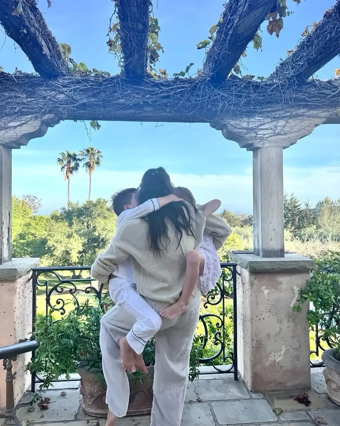 Meghan Markle holds her children, Prince Archie and Princess Lilibet, in an embrace on a balcony beneath a wooden pergola.