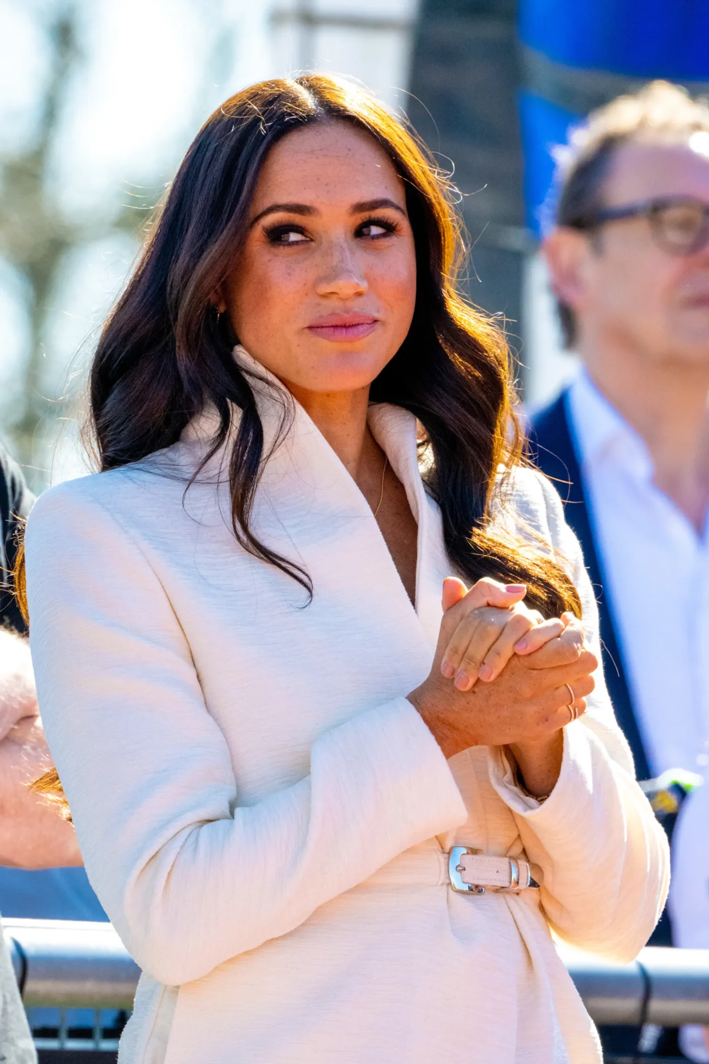 Meghan Markle wearing a white coat with her hands clasped together during a public appearance.