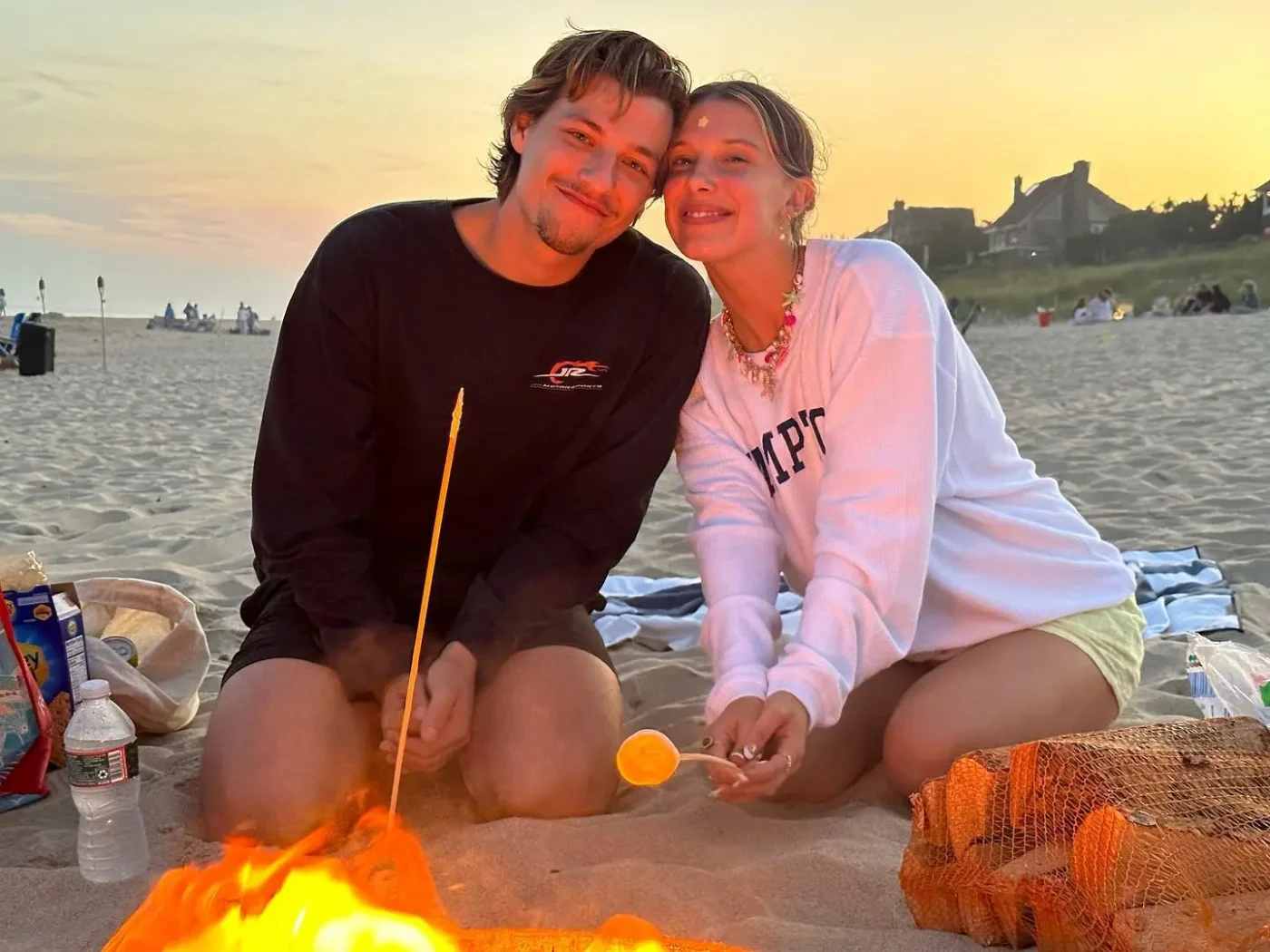 Millie Bobby Brown and Jake Bongiovi sitting by a beach campfire roasting marshmallows.