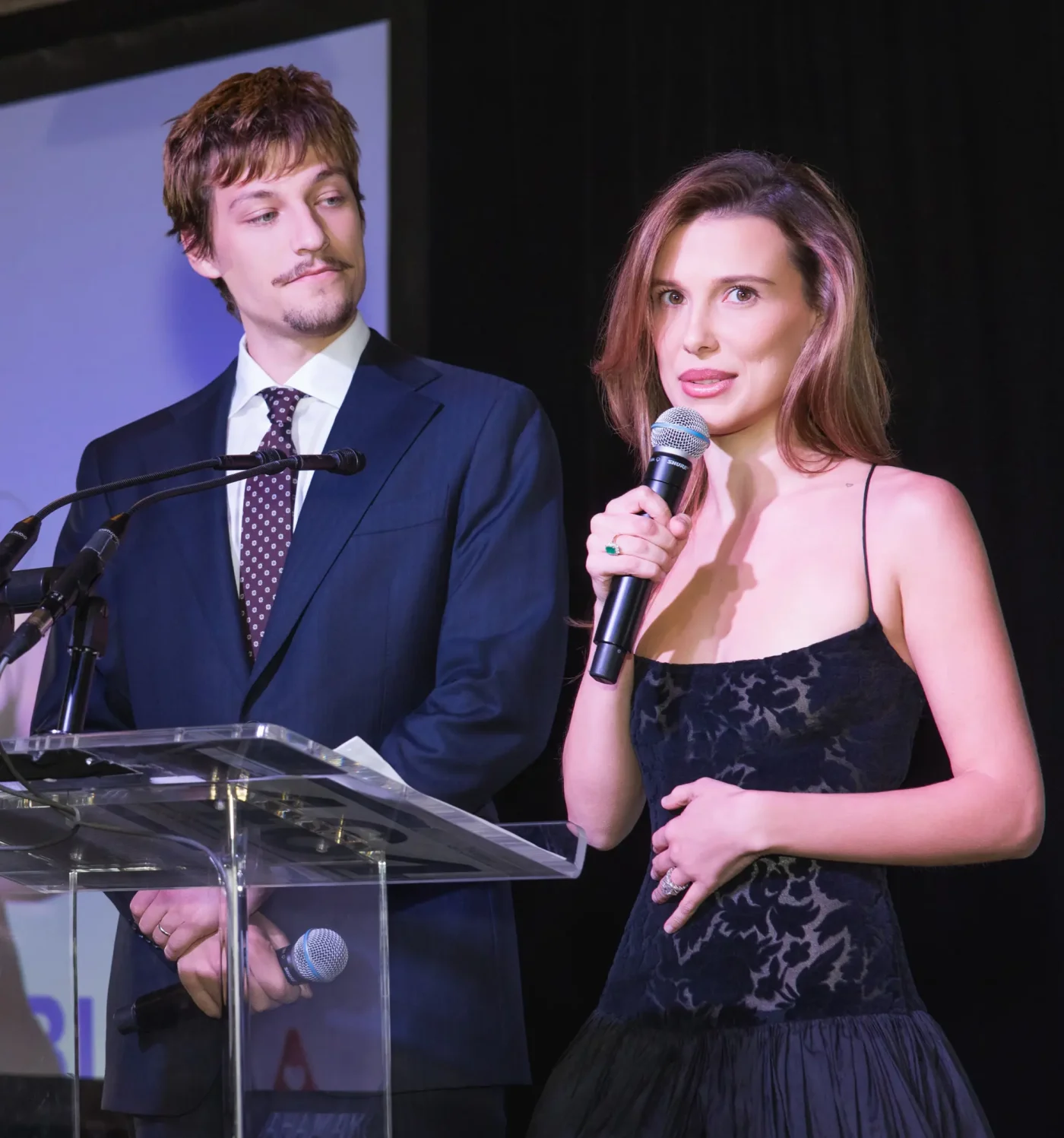 Millie Bobby Brown and Jake Bongiovi speaking at a podium.