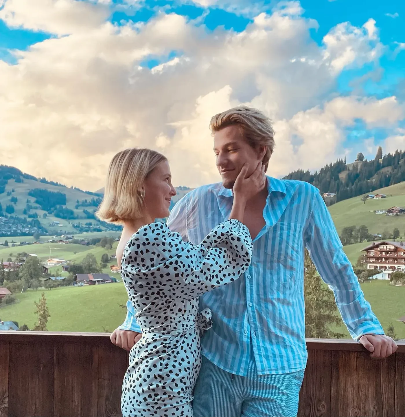 Millie Bobby Brown and Jake Bongiovi standing on a balcony with a scenic mountain landscape in the background.