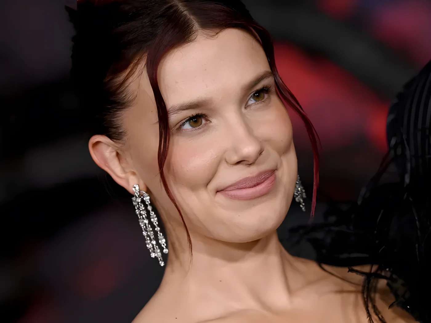 Millie Bobby Brown smiles as she looks off intot he distance. She is wearing diamond chandelier earrings, and her dark hair is styled in an updo with thin, face-framing pieces.