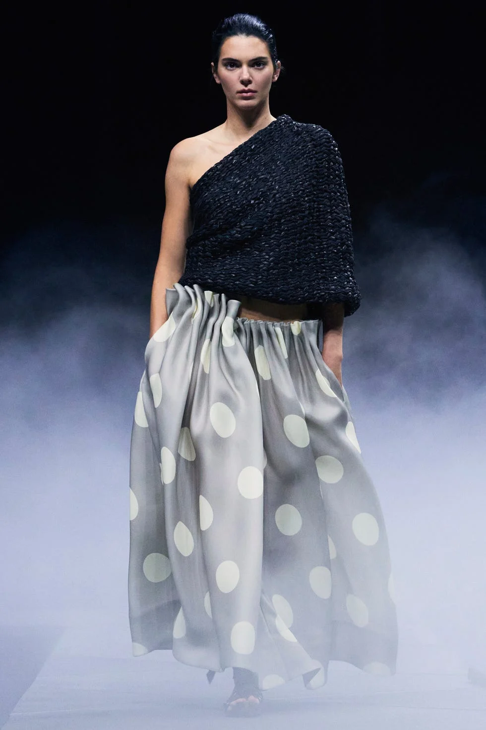 Model wearing a one-shoulder black top and a polka dot skirt on a foggy runway.
