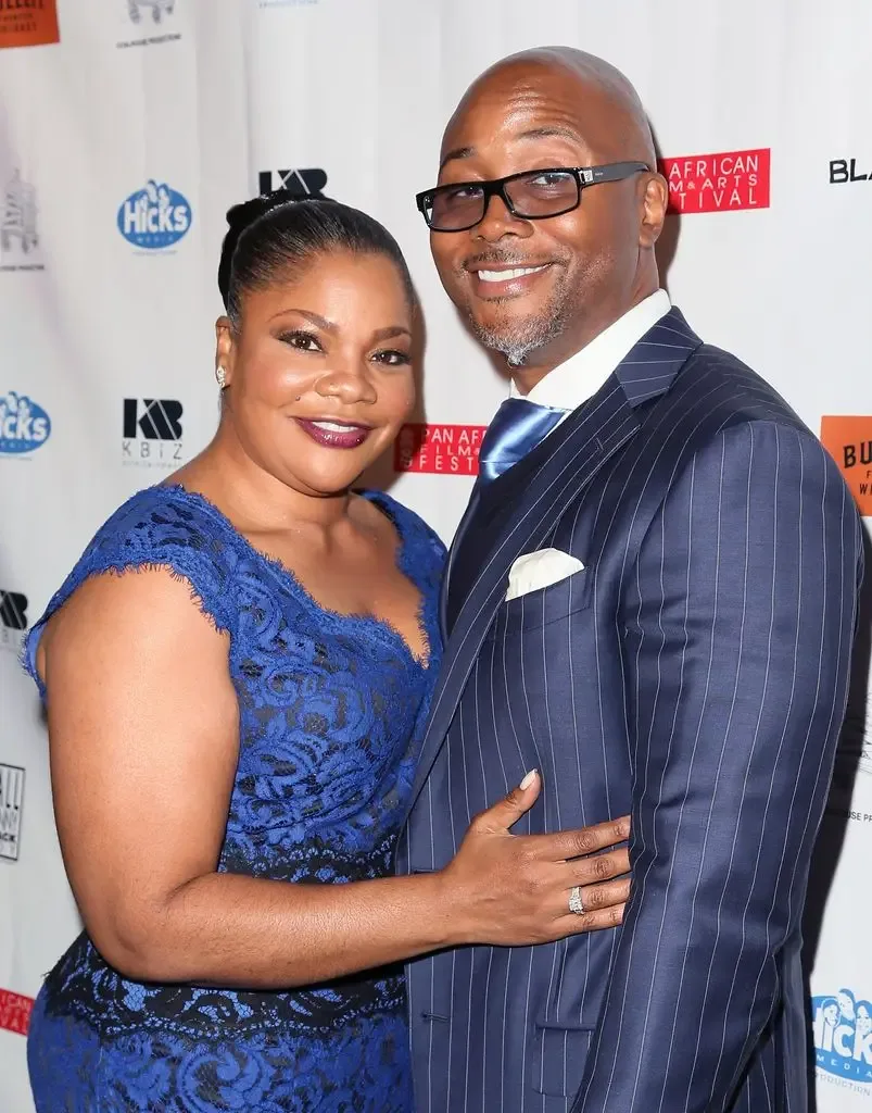 Mo'Nique in a blue lace dress stands with her arm around husband Sidney Hicks, who wears a pinstripe suit, both smiling at a red carpet event.