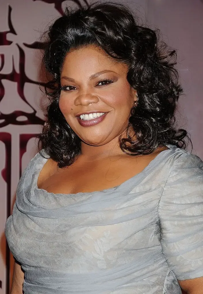 Mo'Nique smiles for photographers, wearing a grey draped off-the-shoulder dress with her dark curly hair styled loosely around her shoulders.