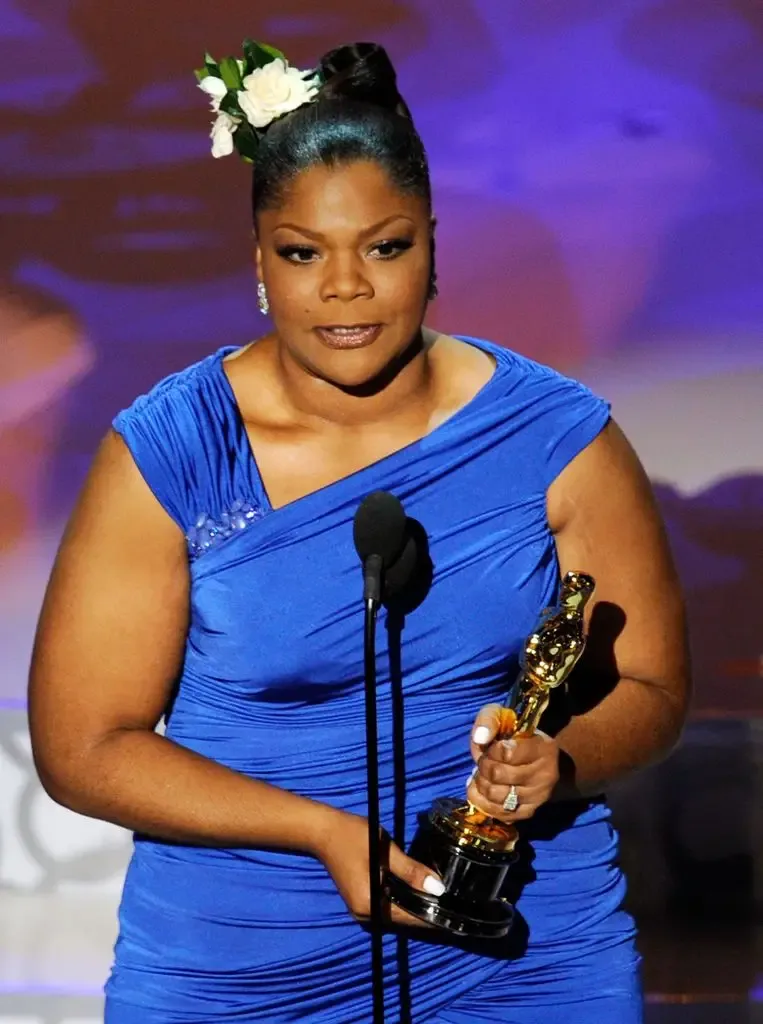 Mo'Nique, wearing a blue gown and a white flower in her hair, speaks at the microphone while holding her Oscar statuette.
