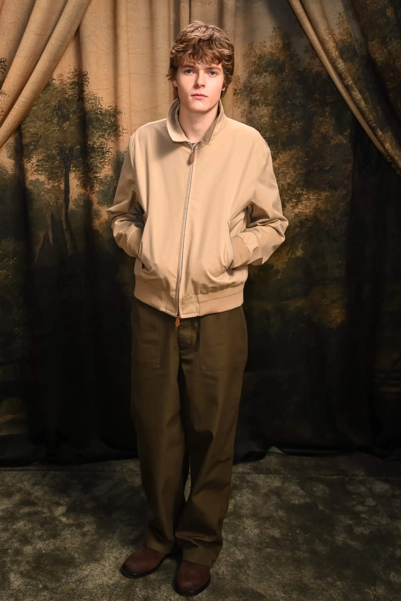 Moses Martin stands in front of a painted backdrop wearing a light beige jacket and dark trousers.
