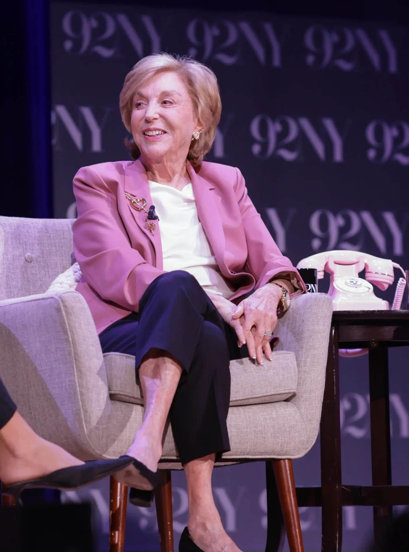 Nora Ephron sitting in a chair onstage at 92NY, wearing a pink blazer.