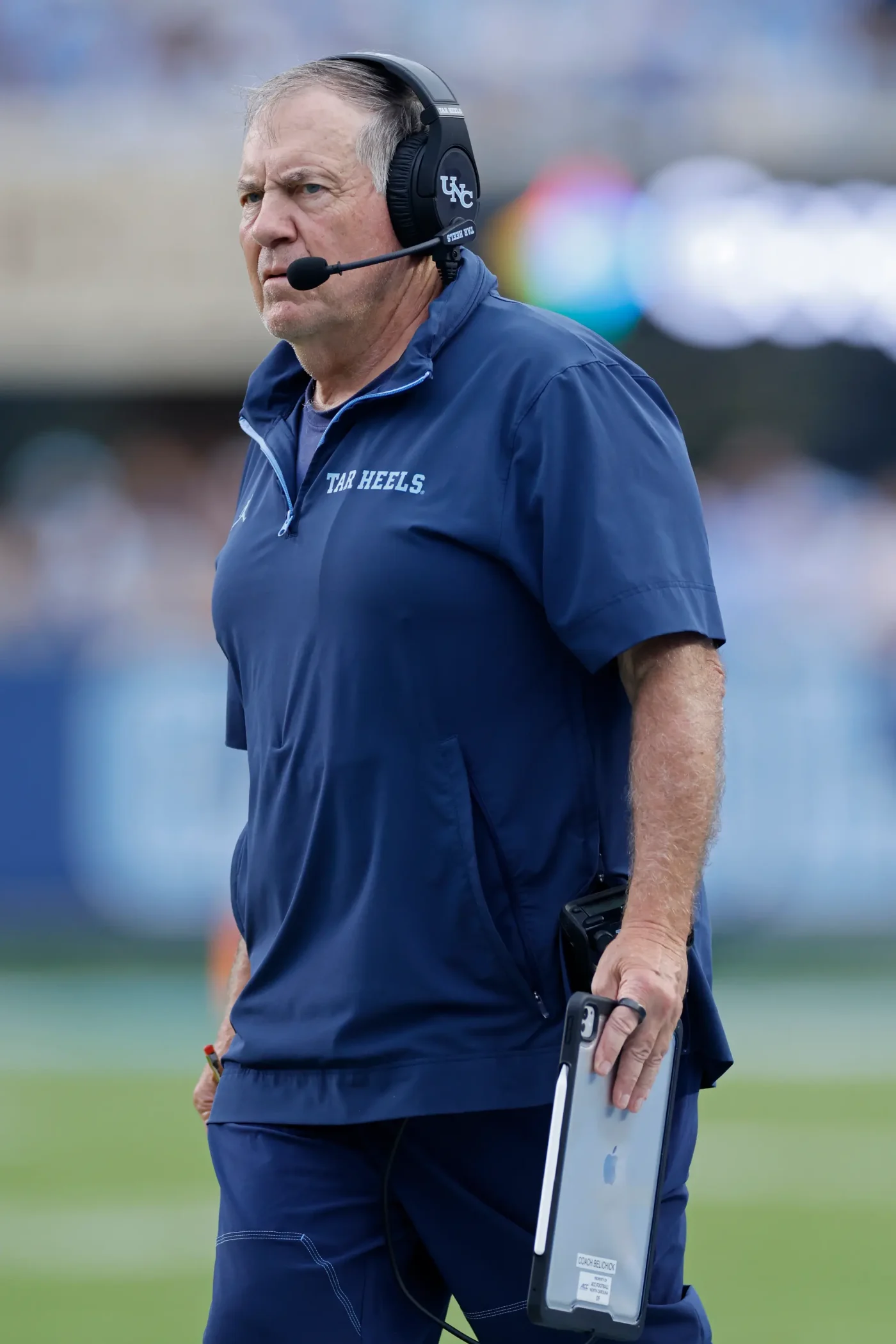 North Carolina Tar Heels head coach Bill Belichick wearing a headset and navy blue pullover while holding a tablet.