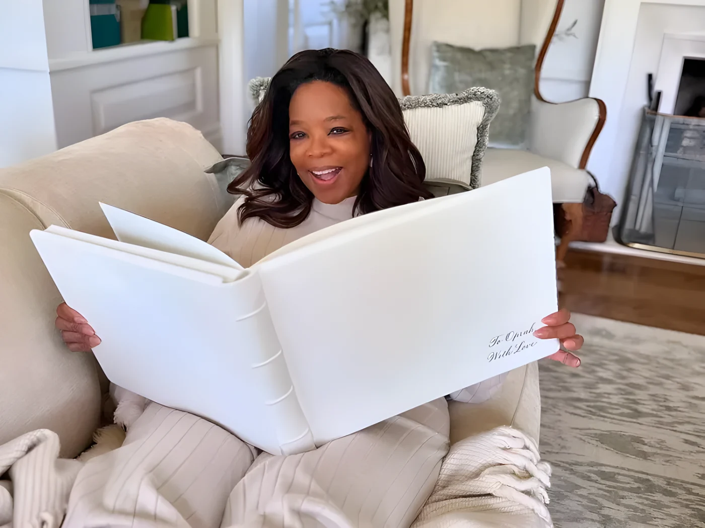 Oprah Winfrey holding a large open book while seated in a cozy living room setting