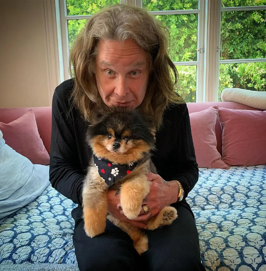 Ozzy Osbourne holds a small fluffy dog wearing a patterned bandana while sitting on a couch.