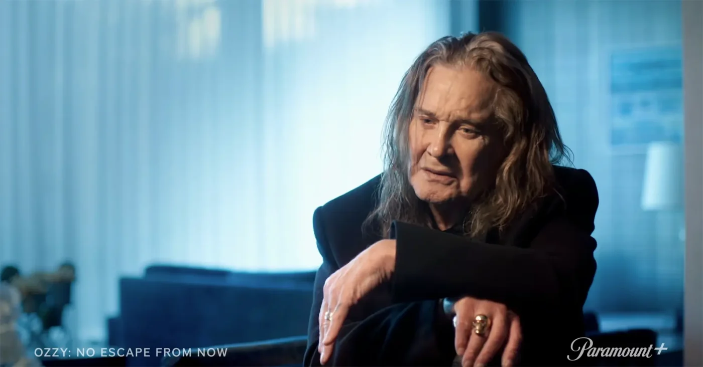 Ozzy Osbourne wearing a black jacket, seated with hands clasped in front of him.
