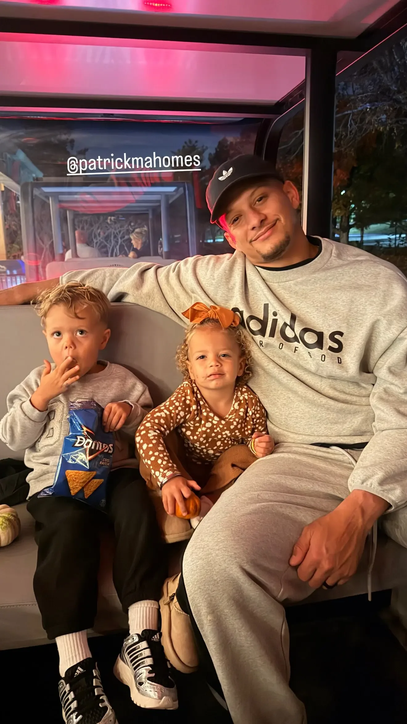 Patrick Mahomes sits in a vehicle with two of his children, a boy eating chips and a young girl holding a small pumpkin.