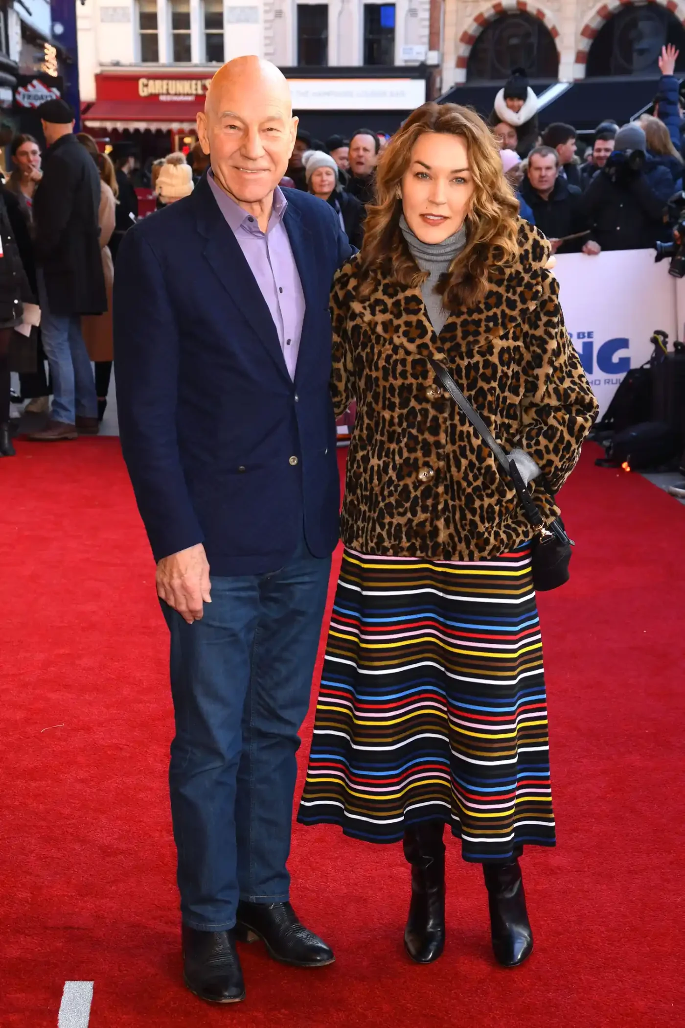Patrick Stewart and his daughter Sophie Stewart smile as they pose side by side on the red carpet. Stewart is wearing a dark blue suit jacket over a light purple dress shirt and dark trousers. Sophie is wearing an animal-print coat over a colorful striped skirt.