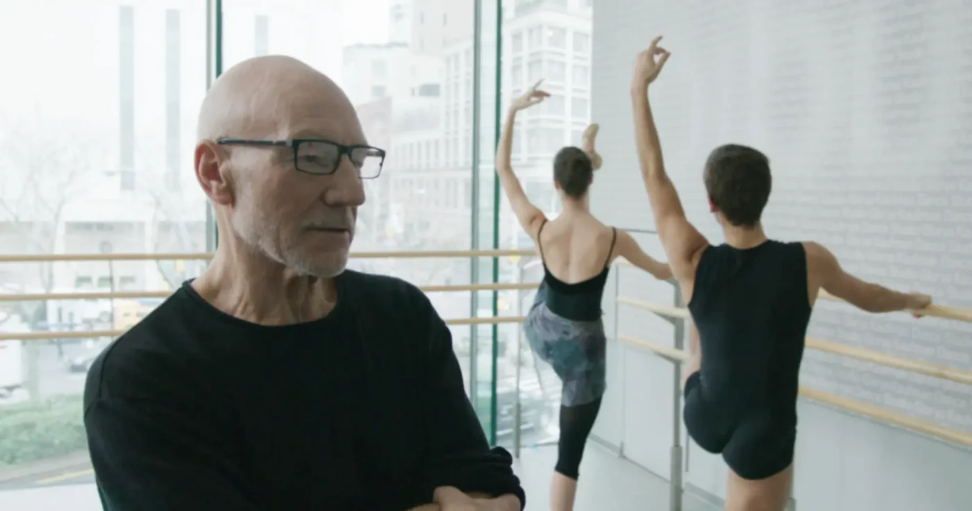 Patrick Stewart in a black shirt and glasses in the foreground with two dancers in a dance studio in the background.