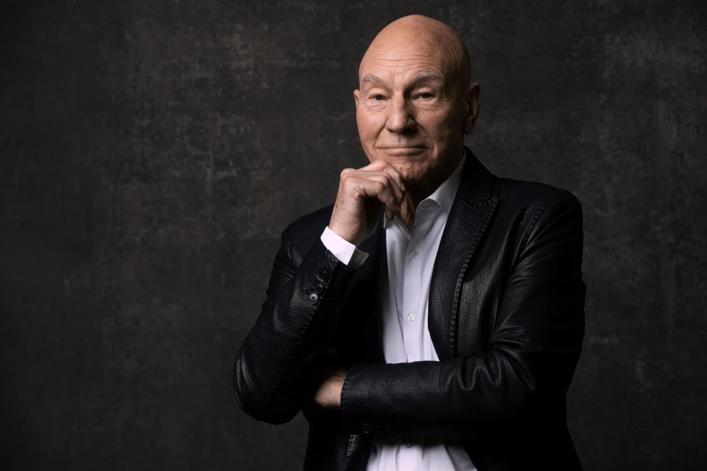 Patrick Stewart poses for a portrait wearing a black leather jacket over a white shirt against a dark textured background.