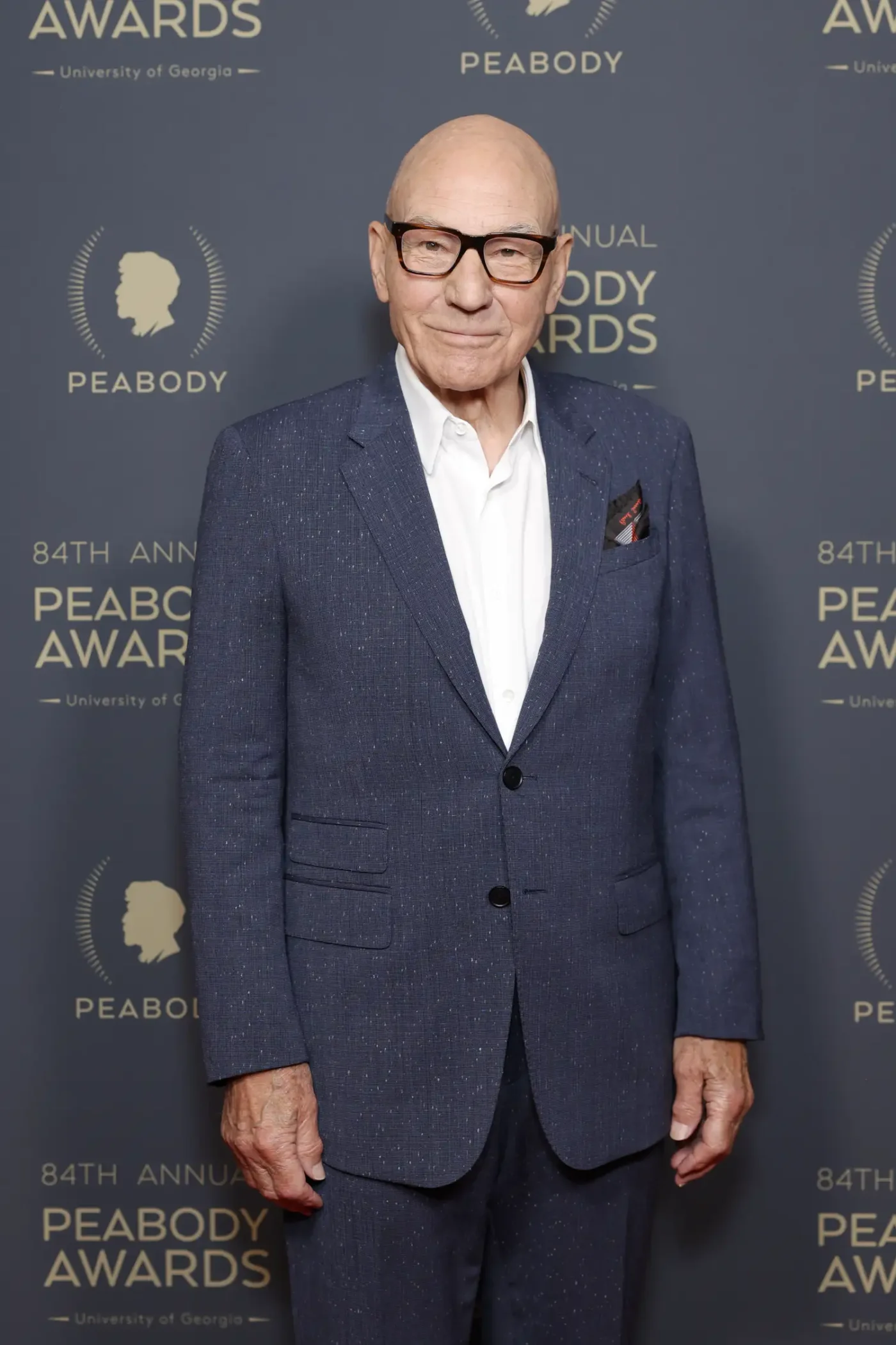 Patrick Stewart smiles as he poses in a dark blue suit and a white dress shirt on the red carpet.