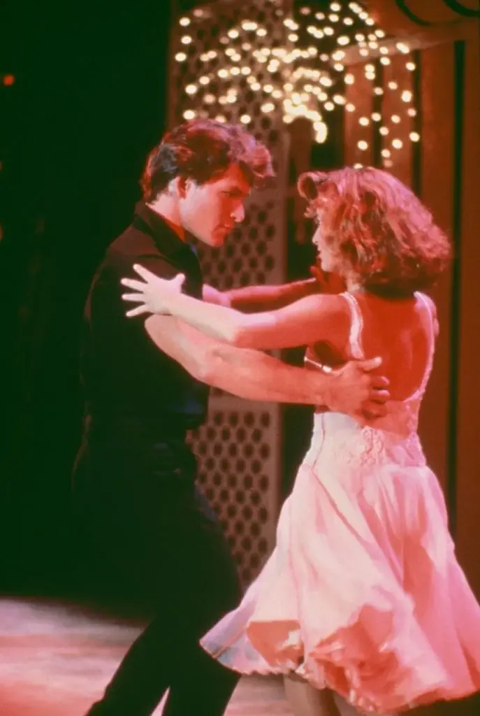Patrick Swayze and Jennifer Grey hold each other in a structured dance pose and lock eyes. Swayze ears a short sleeved black skirt and black trousers while Grey wears a pink dress with a scoop back and full skirt.
