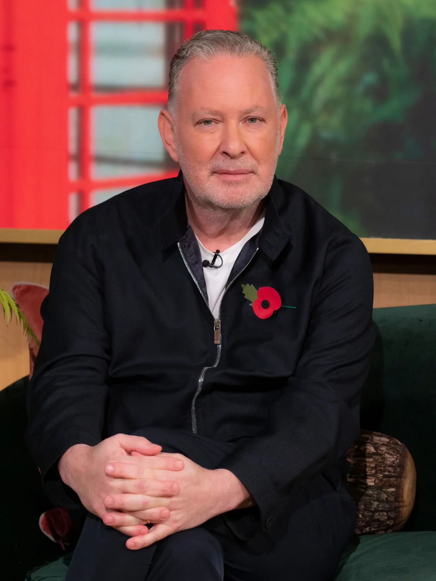 Paul Kemsley sitting on a couch with his hands clasped, wearing a dark jacket with a poppy pinned to the lapel.