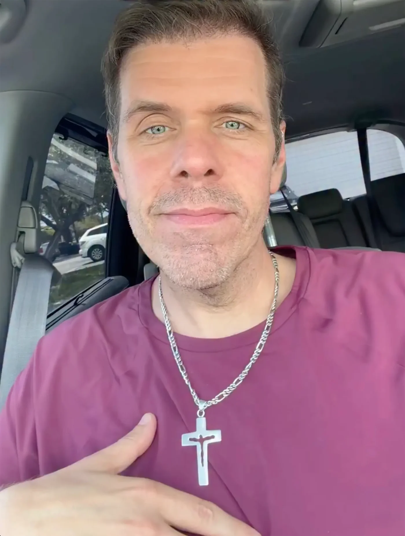 Perez Hilton in a car, wearing a purple shirt and a silver cross necklace.