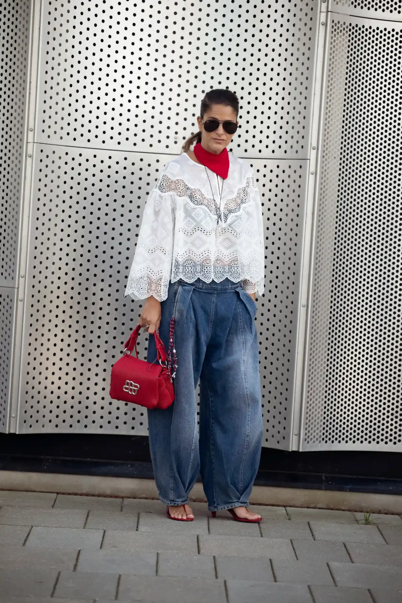 Person in a stylized outfit with a white lace jacket loose denim pants red scarf and handbag posing against a textured background