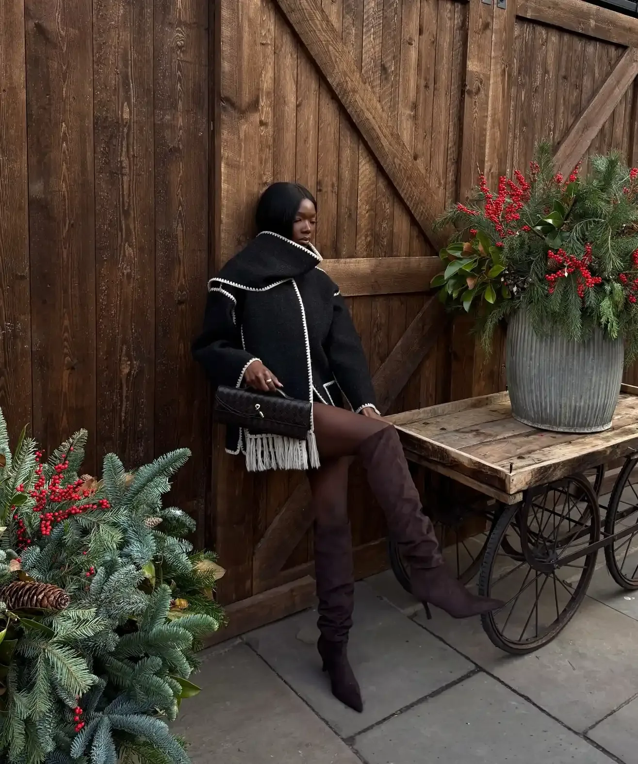 Person leaning against a wooden wall next to a wooden cart adorned with holidaythemed greenery