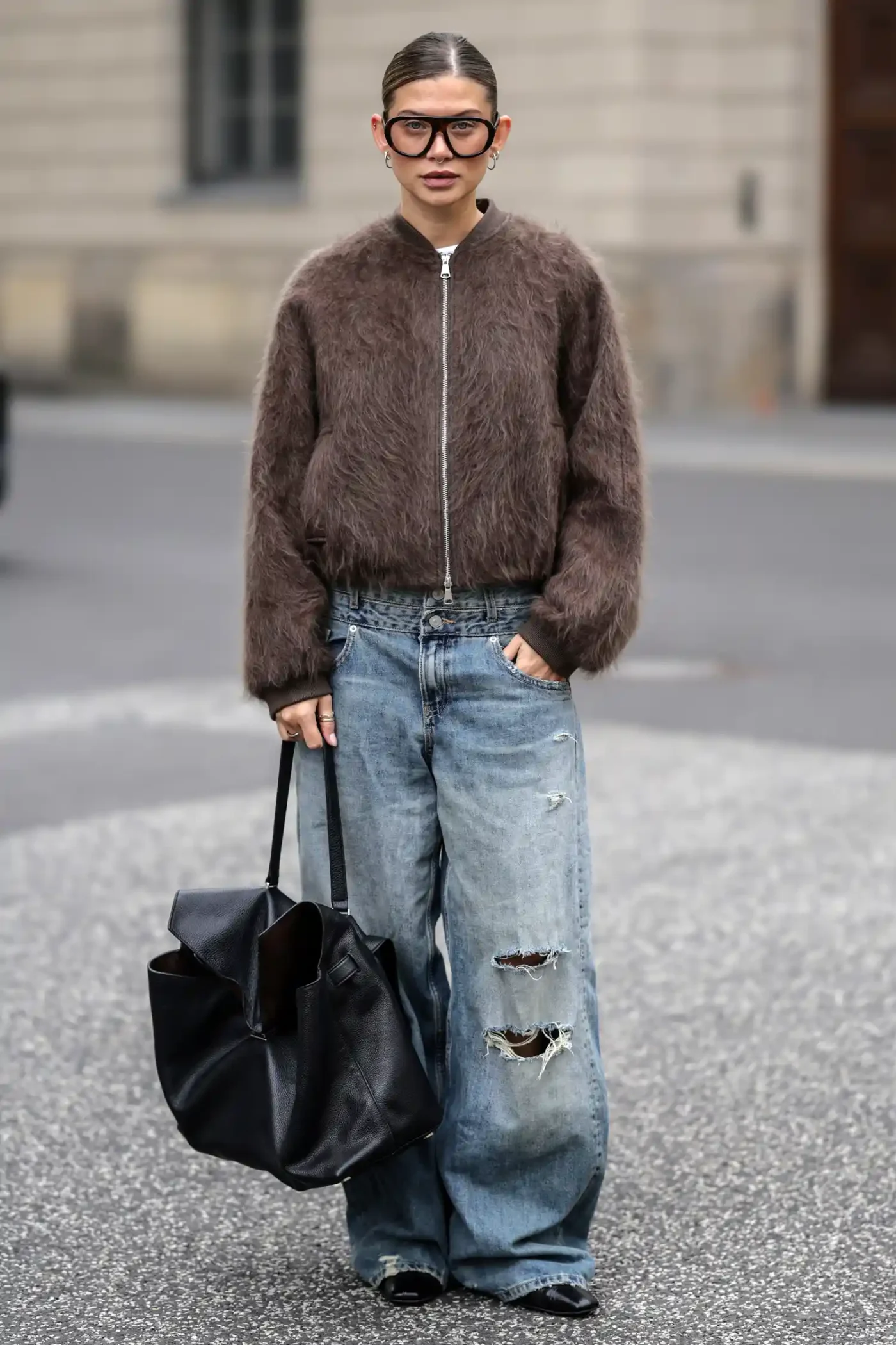 Person wearing a fuzzy jacket ripped jeans and oversize glasses holding a black bag standing on a city street