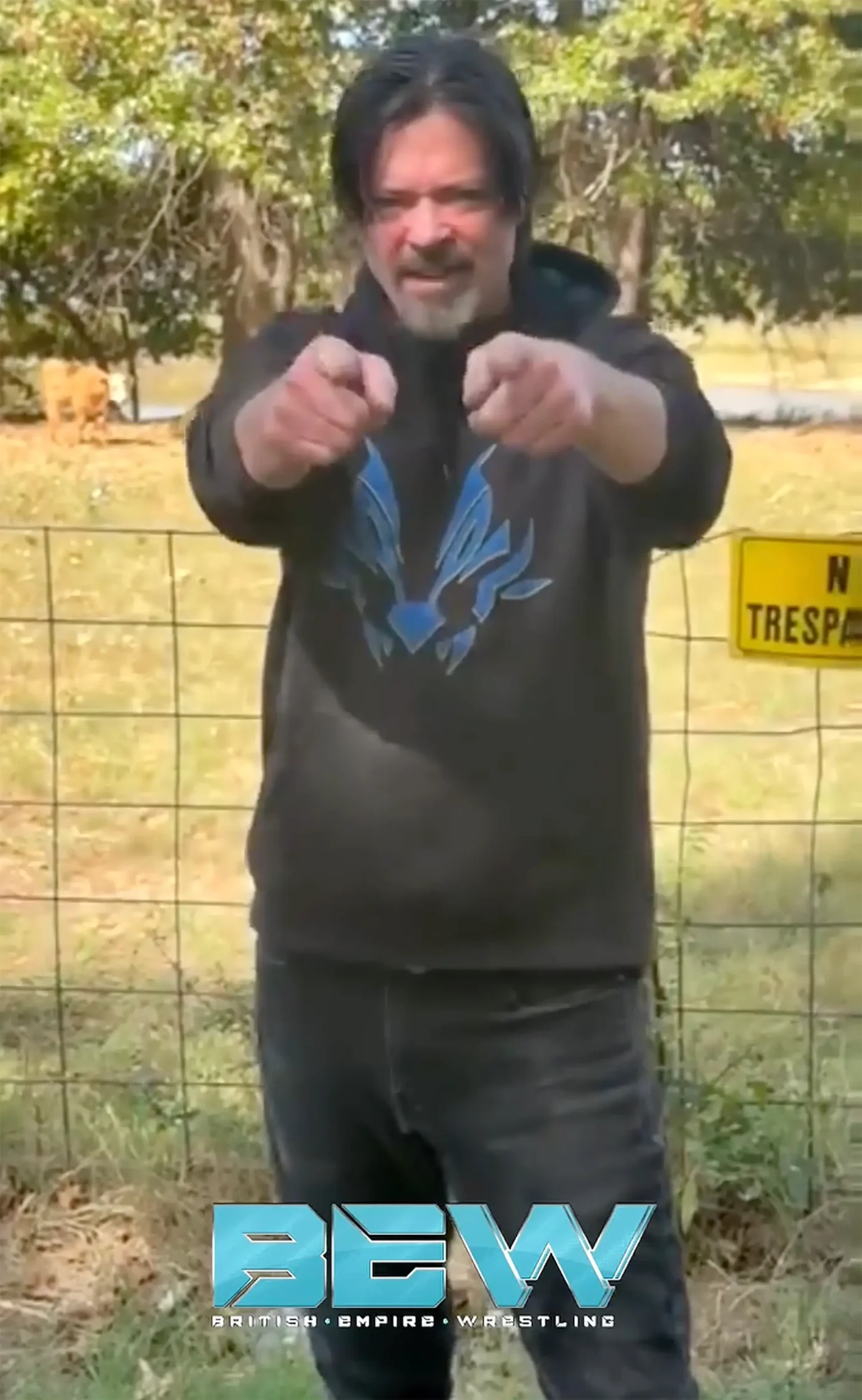 Phillip Andrew points both index fingers forward while wearing a dark hoodie with a blue graphic, standing in front of a wire fence and a grassy field with a