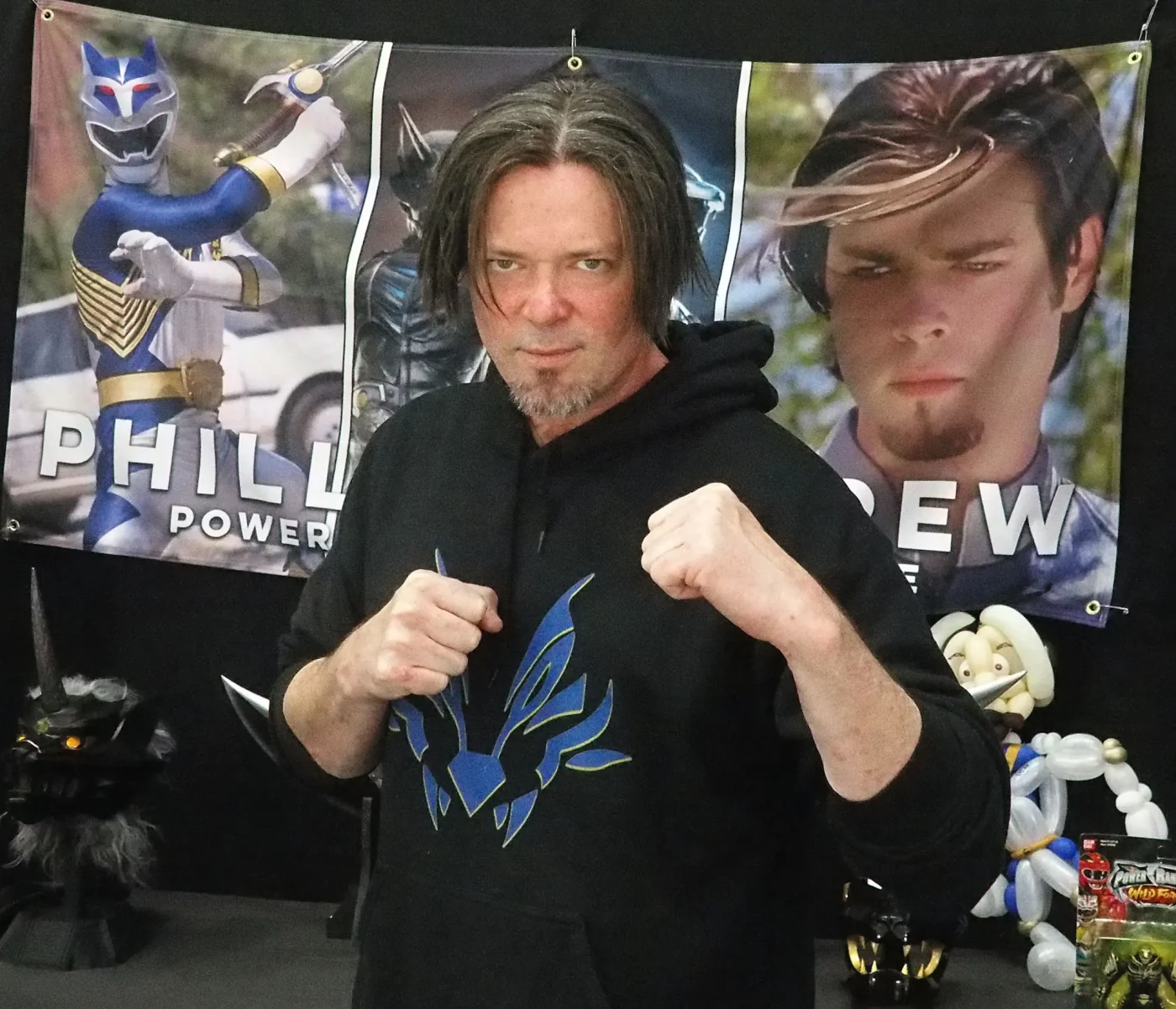 Phillip Andrew poses with his fists raised in front of a banner featuring his character from Power Rangers.