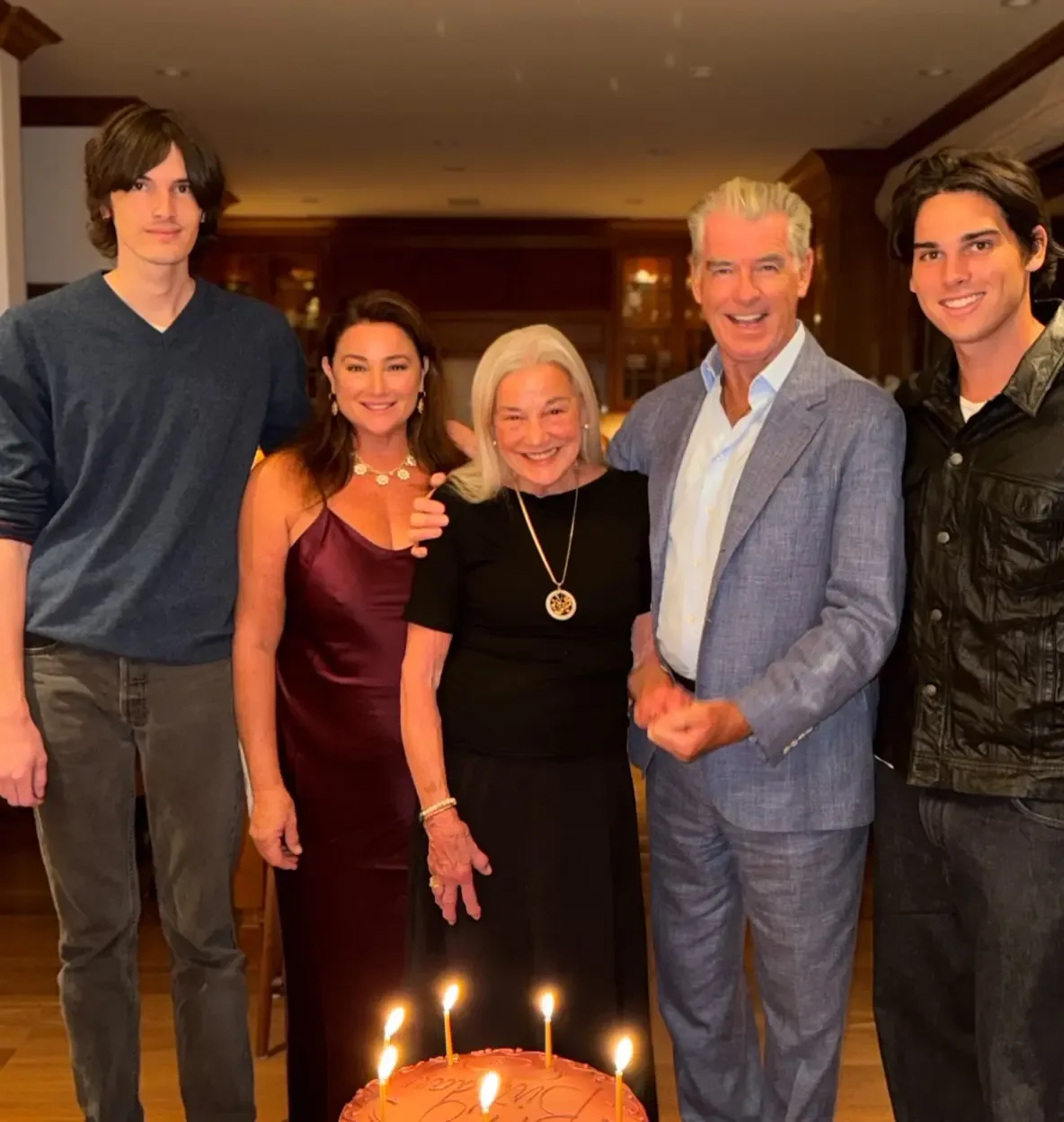 Pierce Brosnan, Keely Shaye Smith, and family members pose behind a birthday cake with lit candles.