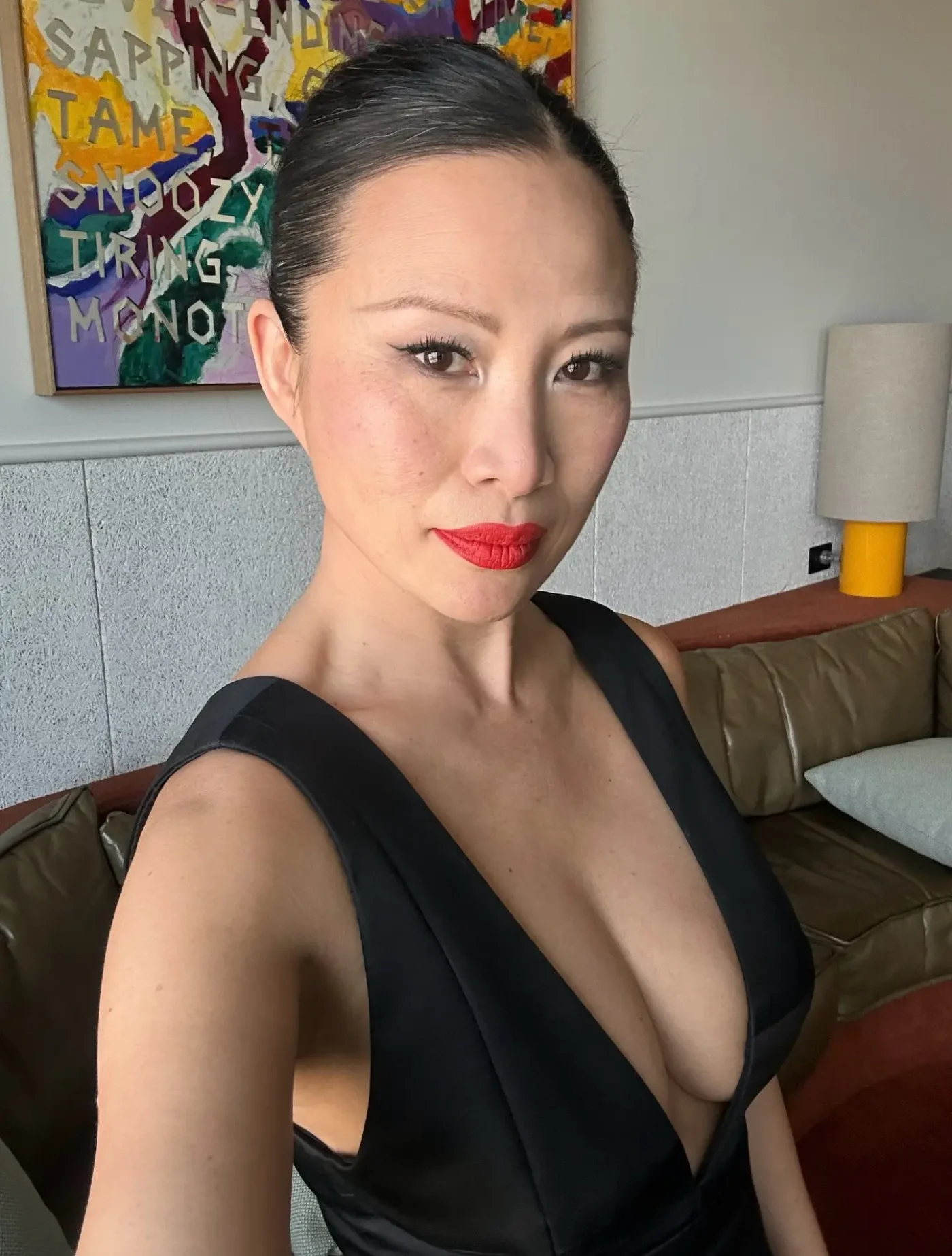 Poh Ling Yeow wearing a black dress with a deep V-neck and bright red lipstick, posing for a selfie in an indoor setting with artwork and a couch visible in the background.
