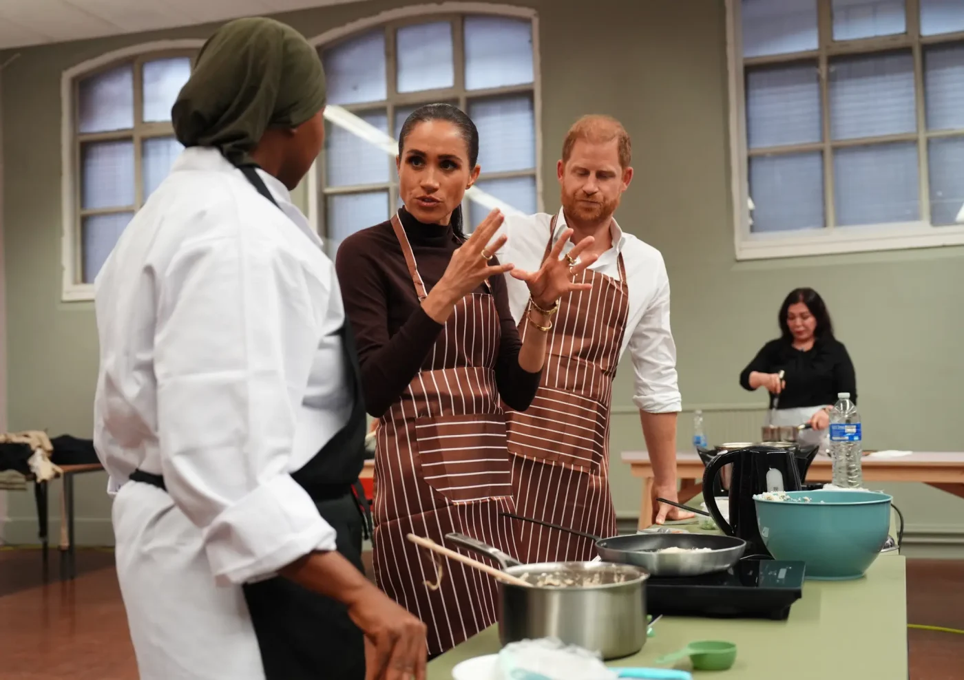 Prince Harry and Meghan, Duchess of Sussex, visit Himmah Hub in Nottingham, wearing matching striped aprons while talking with a chef.