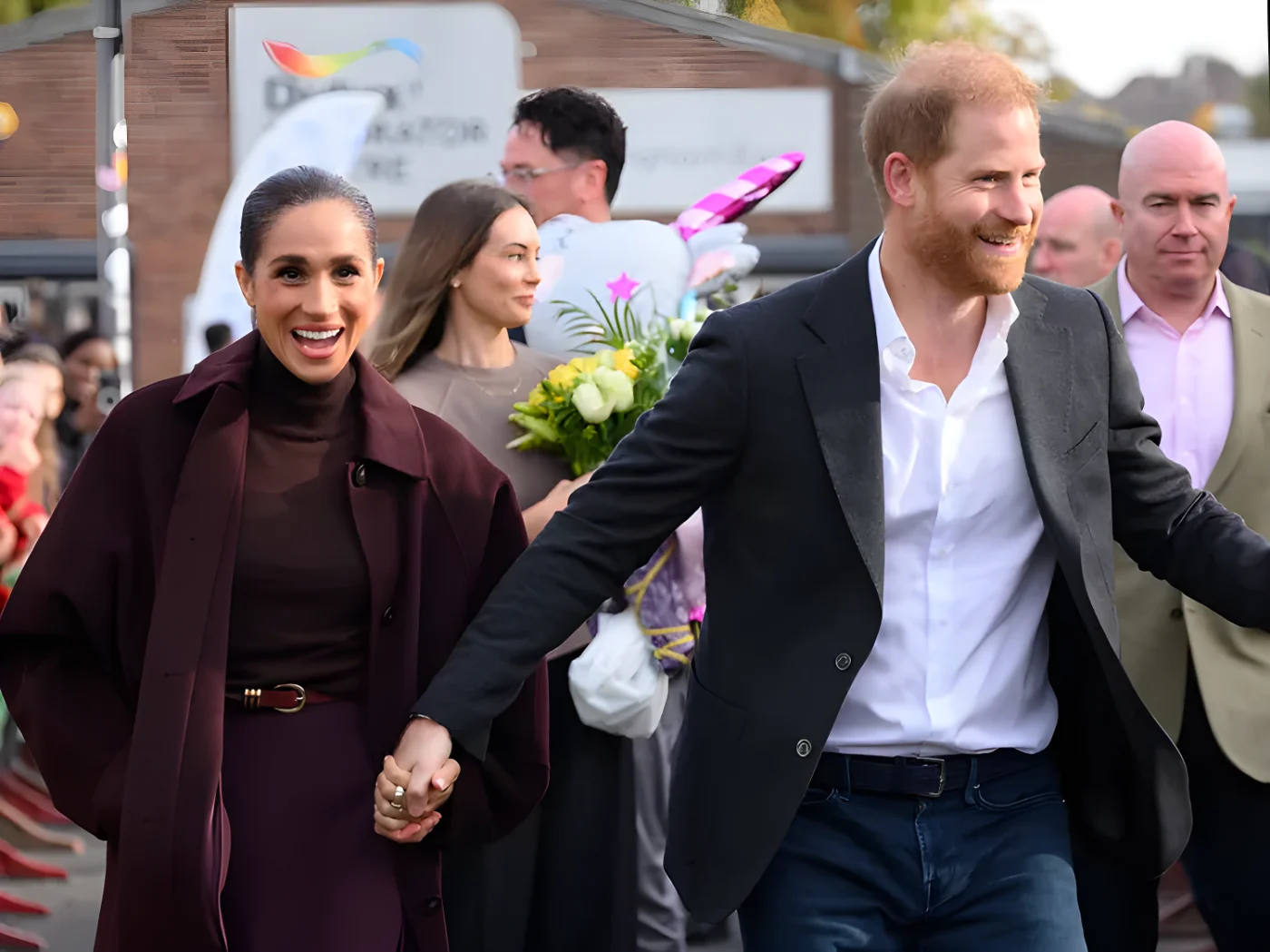 Prince Harry and Meghan Duchess of Sussex walking and holding hands surrounded by other individuals and an outdoor background