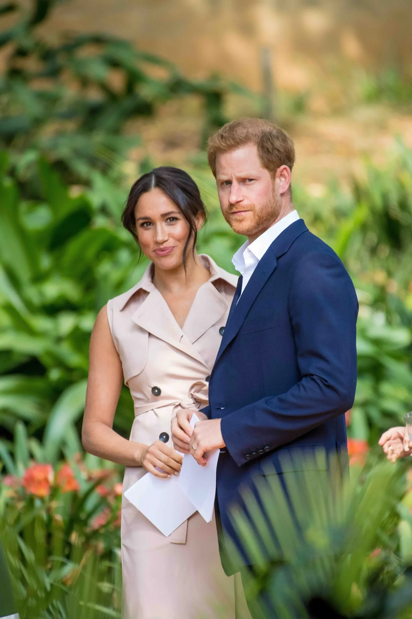 Prince Harry and Meghan Markle standing together outdoors amidst green foliage.