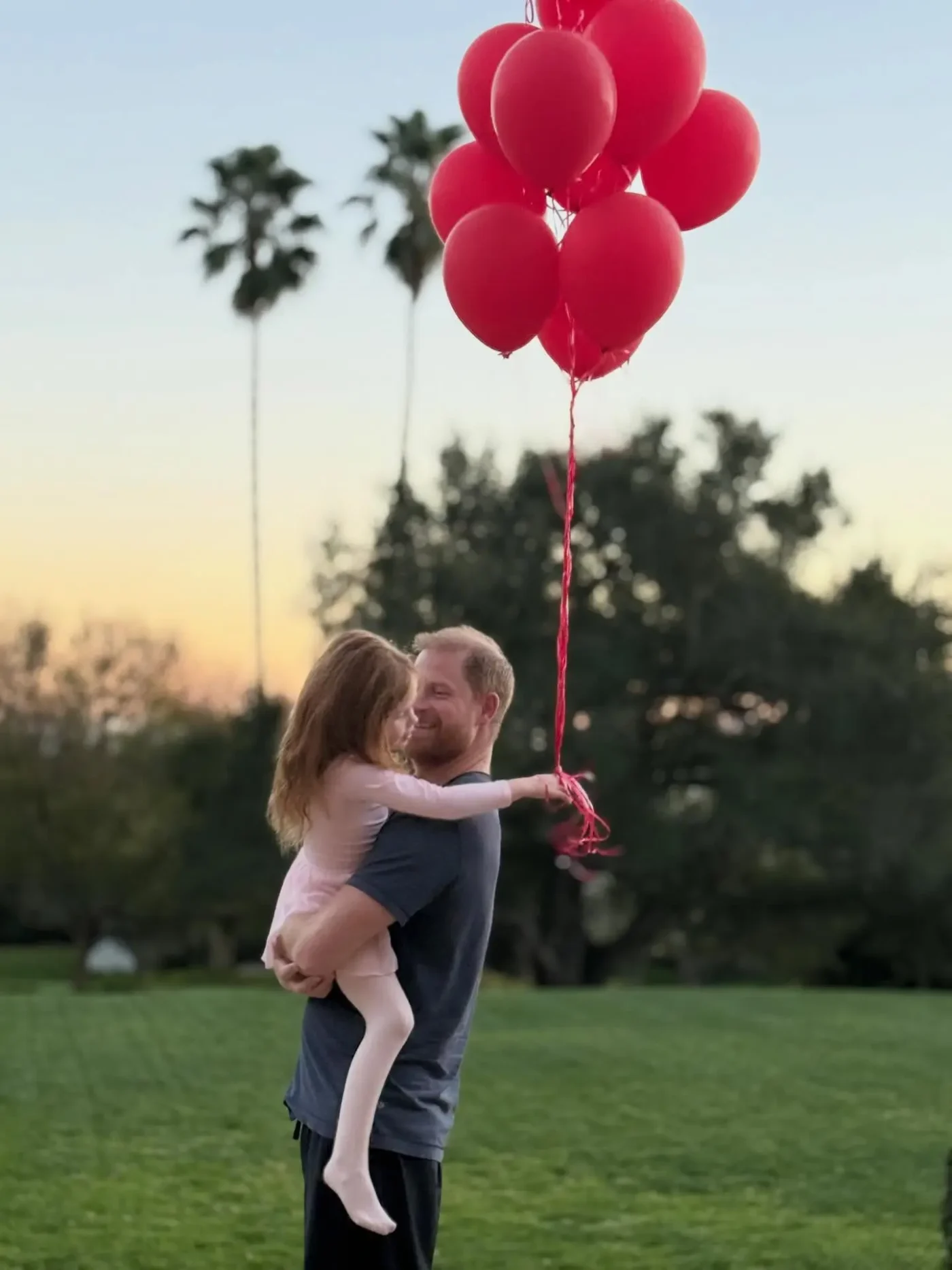 Prince Harry holding Princess Lilibet outdoors with a bunch of red balloons.