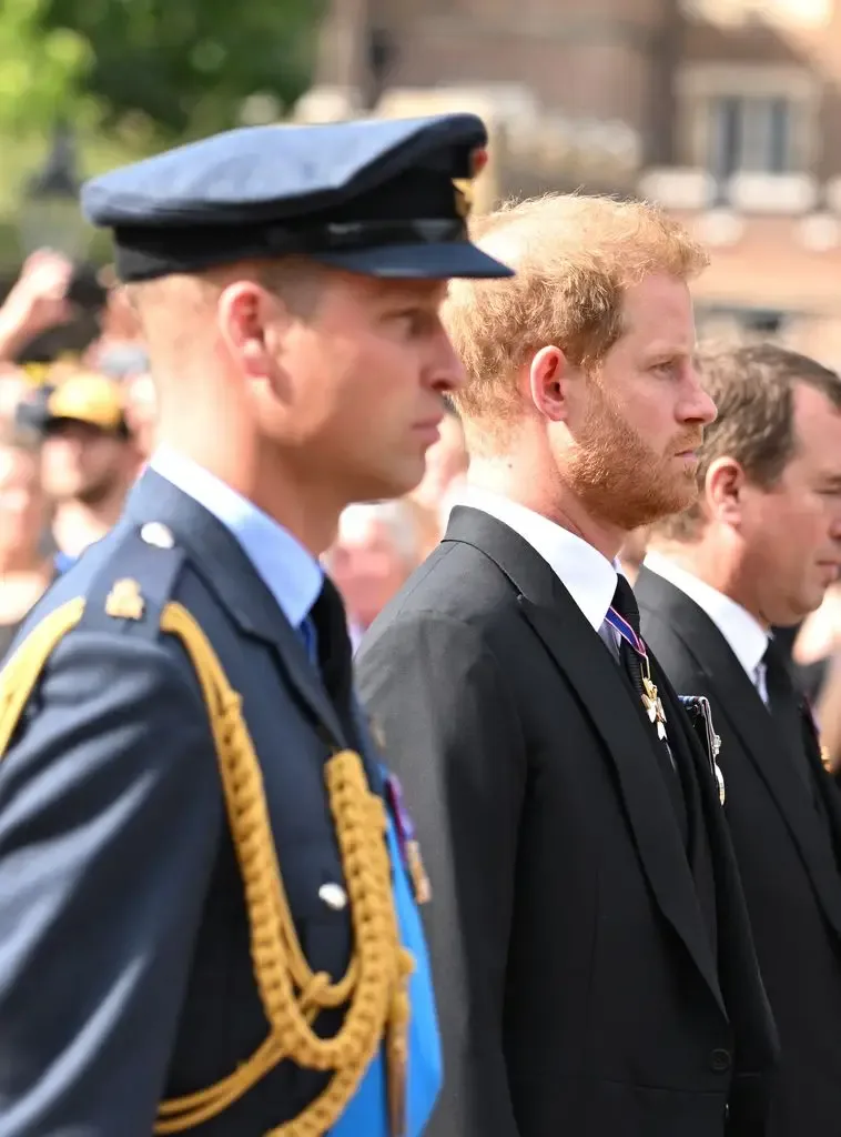 Prince William in military uniform stands next to Prince Harry in a black suit.
