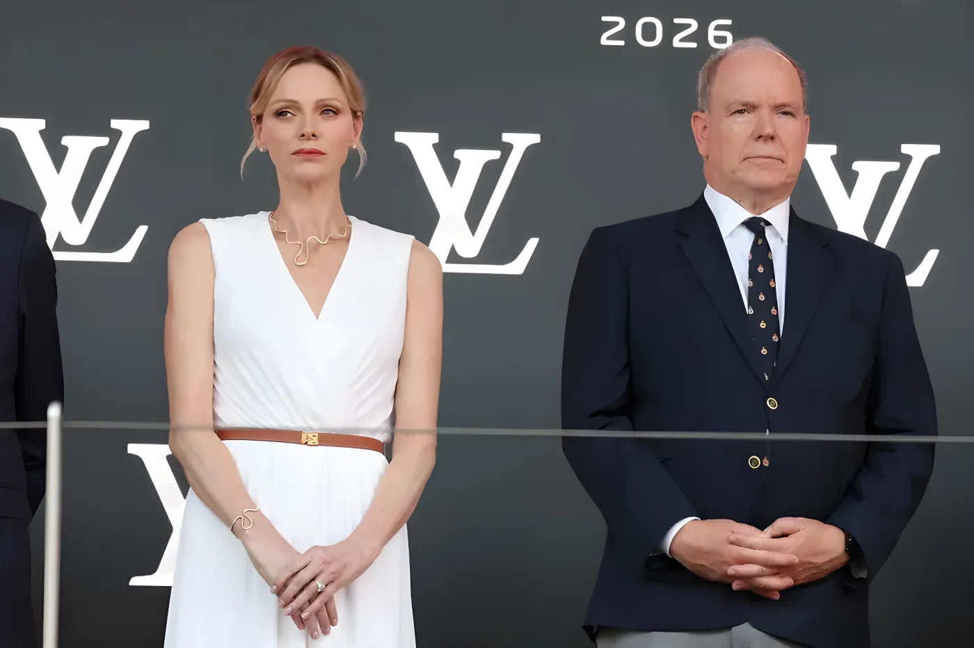 Princess Charlene and Prince Albert stand on a stage with a Louis Vuitton backdrop. The woman wears a white dress and the man a navy suit.