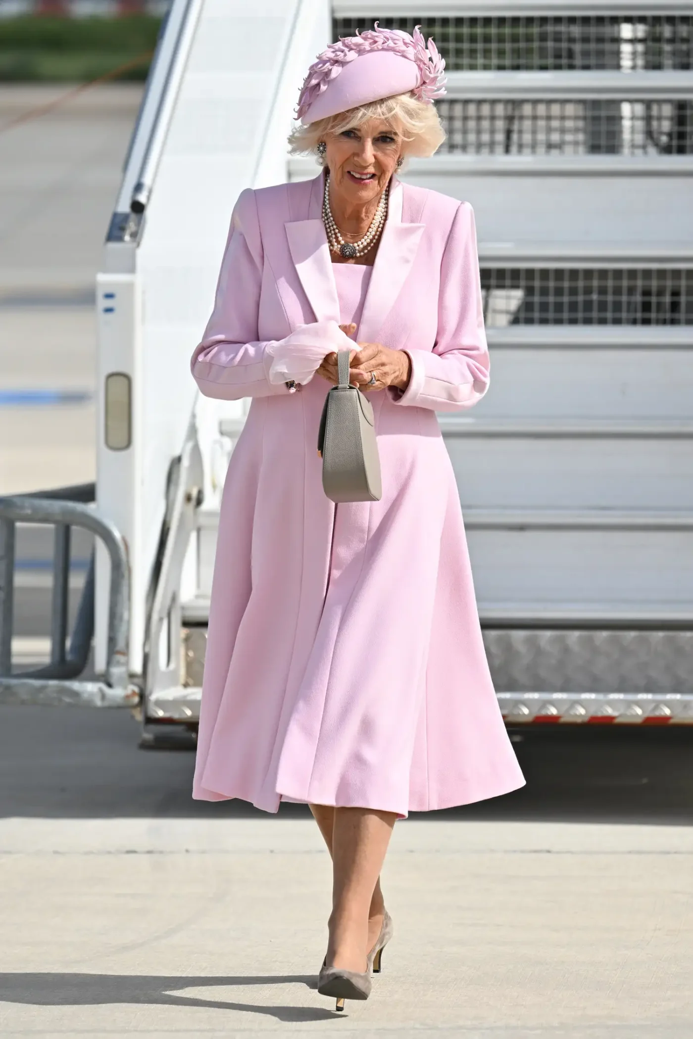 Queen Camilla disembarks a plane while wearing a flared pink trench coat over a matching dress. She styled the look with an intricate hat in light pink, layered pearl necklaces, and suede heels in gray.